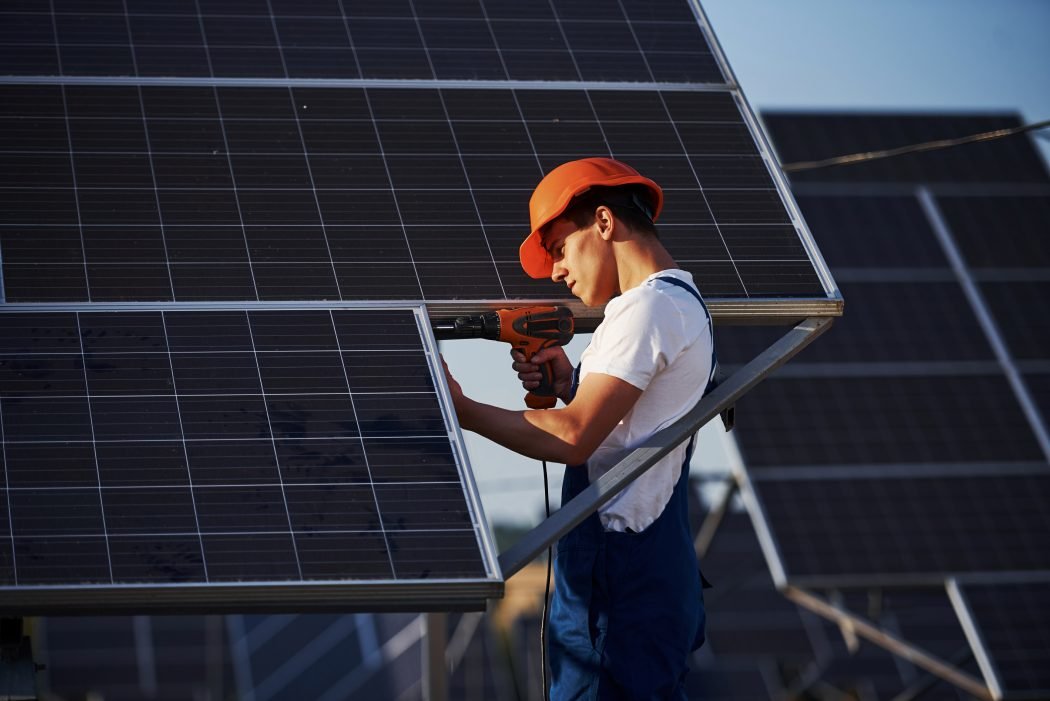 Cordless screwdriver. Male worker in blue uniform outdoors with solar batteries at sunny day